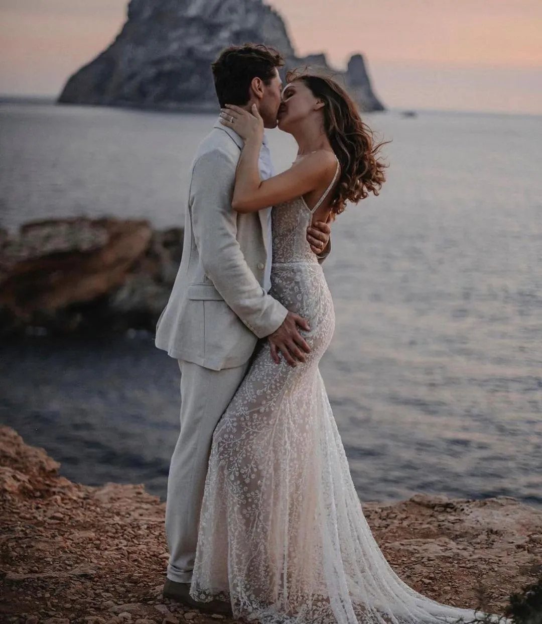 Couple kissing on a rocky beach with the woman wearing a lace wedding dress.