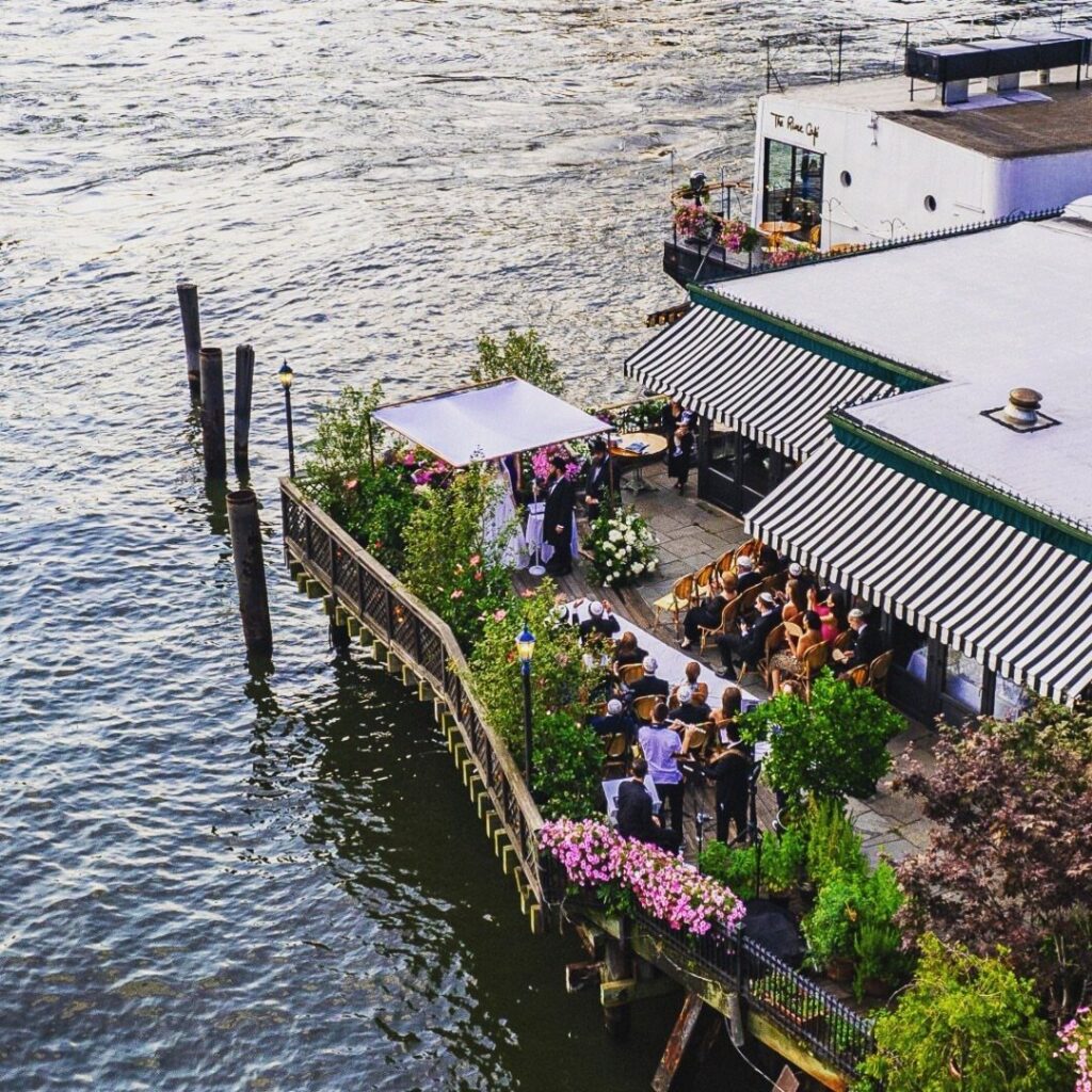River Cafe waterfront patio with guests dining under striped awnings by the river.