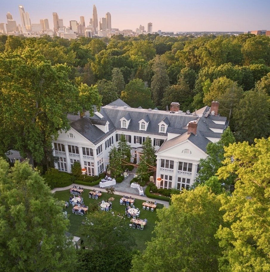 White historic mansion with manicured gardens and event tables set against a city skyline.