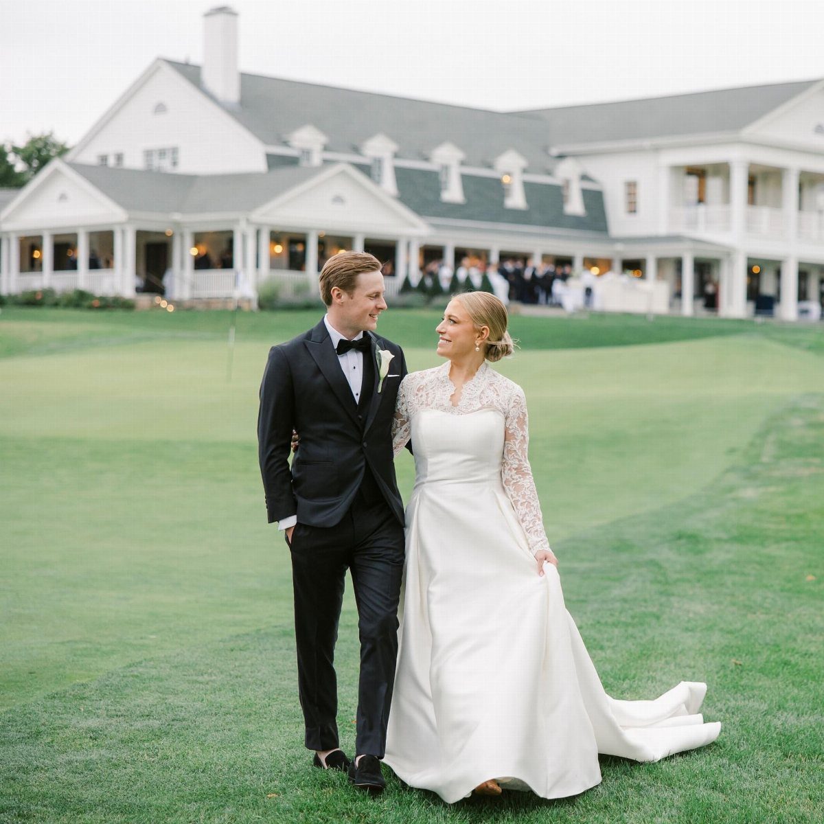 Bride and groom posing on a green lawn before a white wedding venue.
