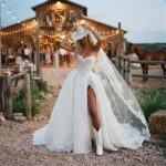 Bride in a white wedding dress, cowboy hat, and boots at a rustic barn.