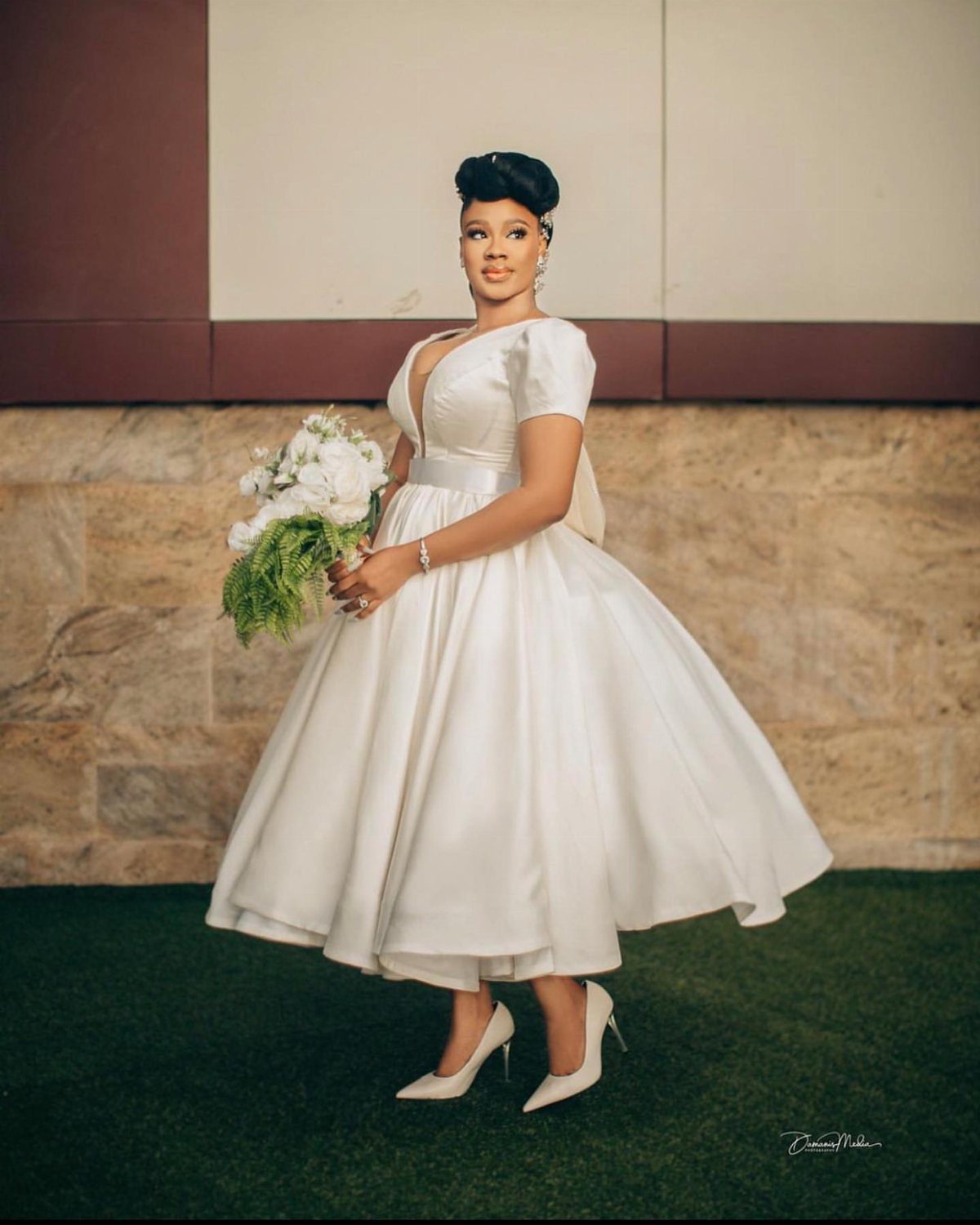 Woman in a white tea-length satin wedding dress holding a white floral bouquet.