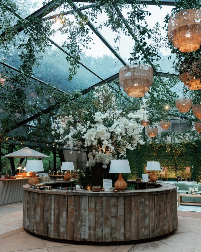 Circular wooden bar with white flowers under a glass roof with hanging lights.