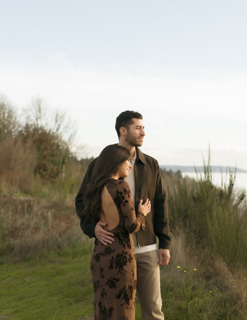 Couple in brown autumn clothing embracing in a grassy field by the water