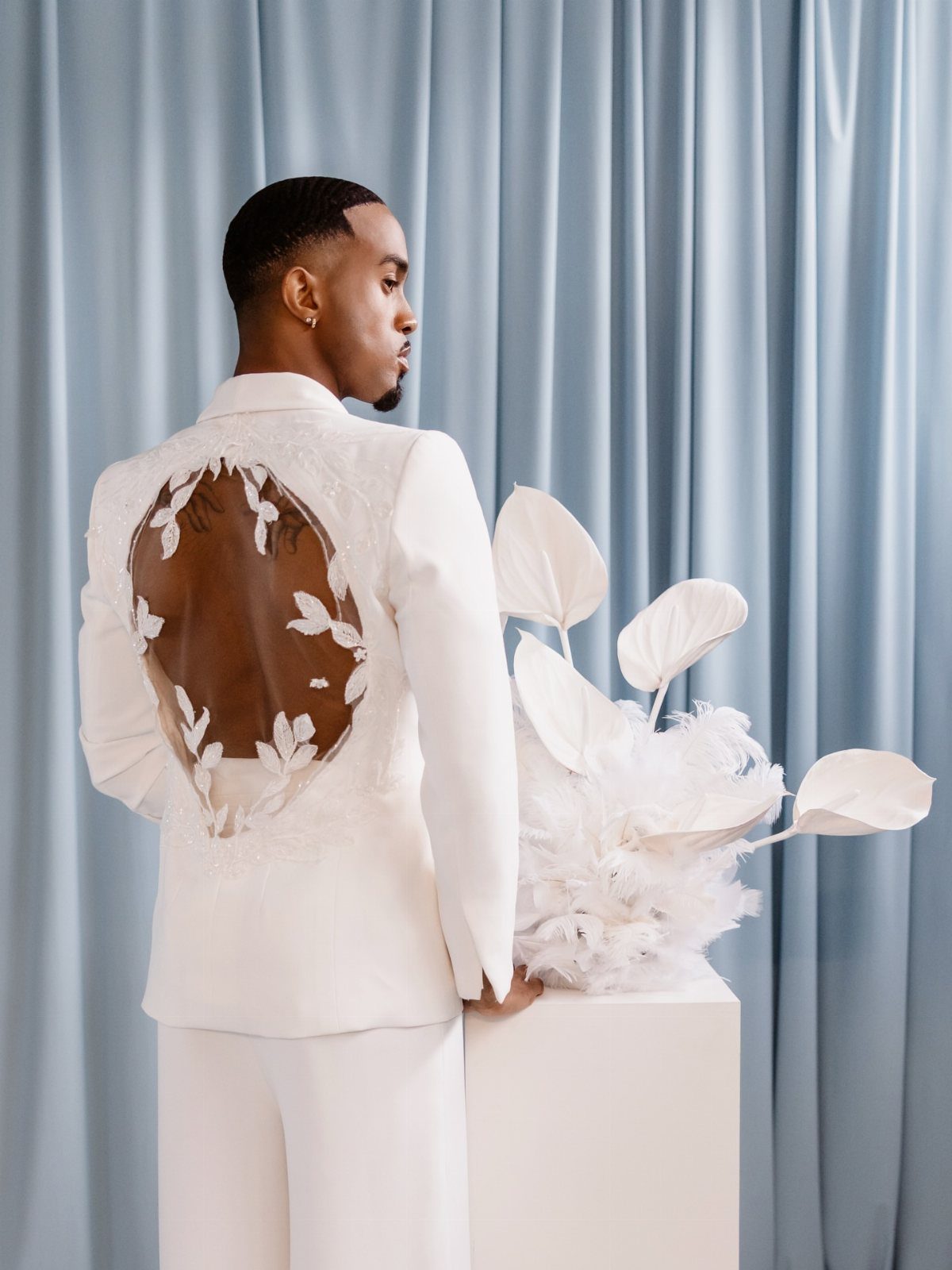 Man in a white suit with an embroidered sheer back panel against blue curtains.
