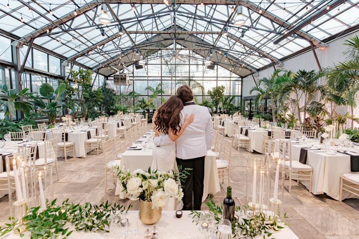 Couple standing in a glass greenhouse wedding venue with white tables and tropical plants