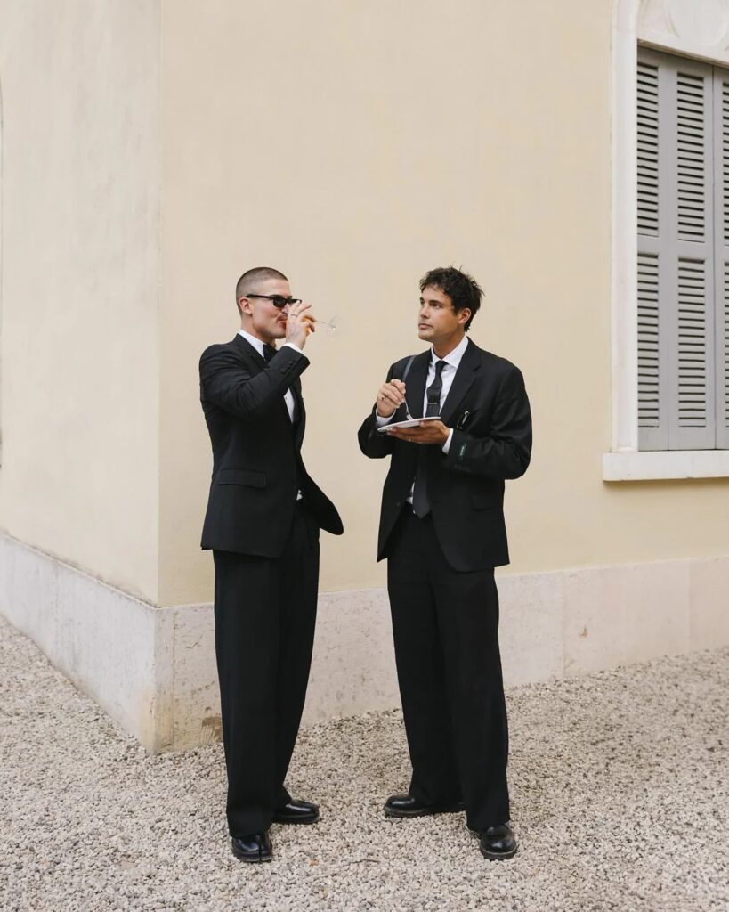 Two men in formal black suits and ties standing outdoors by a wall.