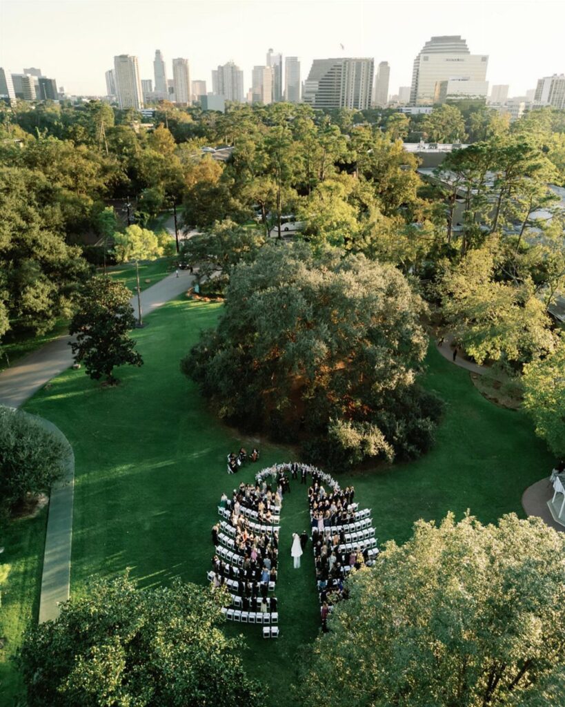 Wedding ceremony guests seated on a green lawn under a massive oak tree.