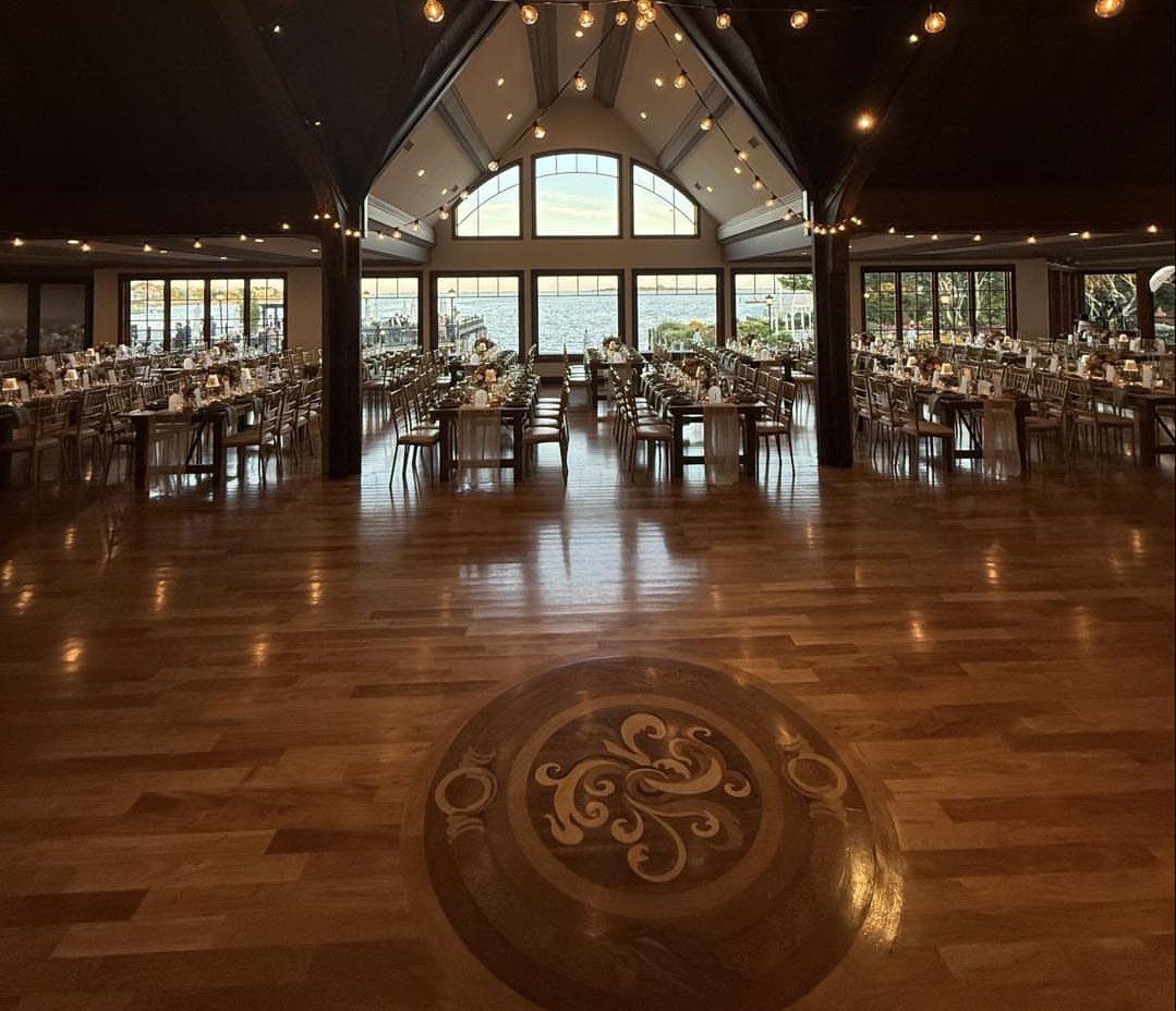 Wedding reception hall with polished wood floors, set tables, and waterfront views.