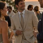 Man in a beige linen suit and tie at a beach wedding sunset.