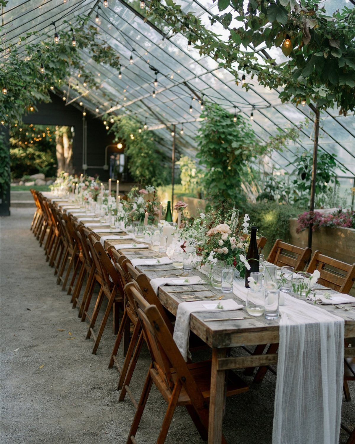 Long wooden wedding table with floral centerpieces inside a bright glass greenhouse.