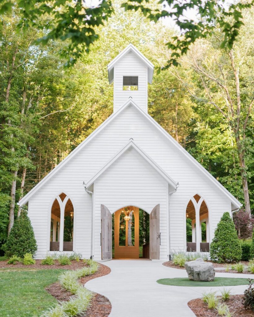 White wooden chapel with a steeple surrounded by green trees and a paved path.