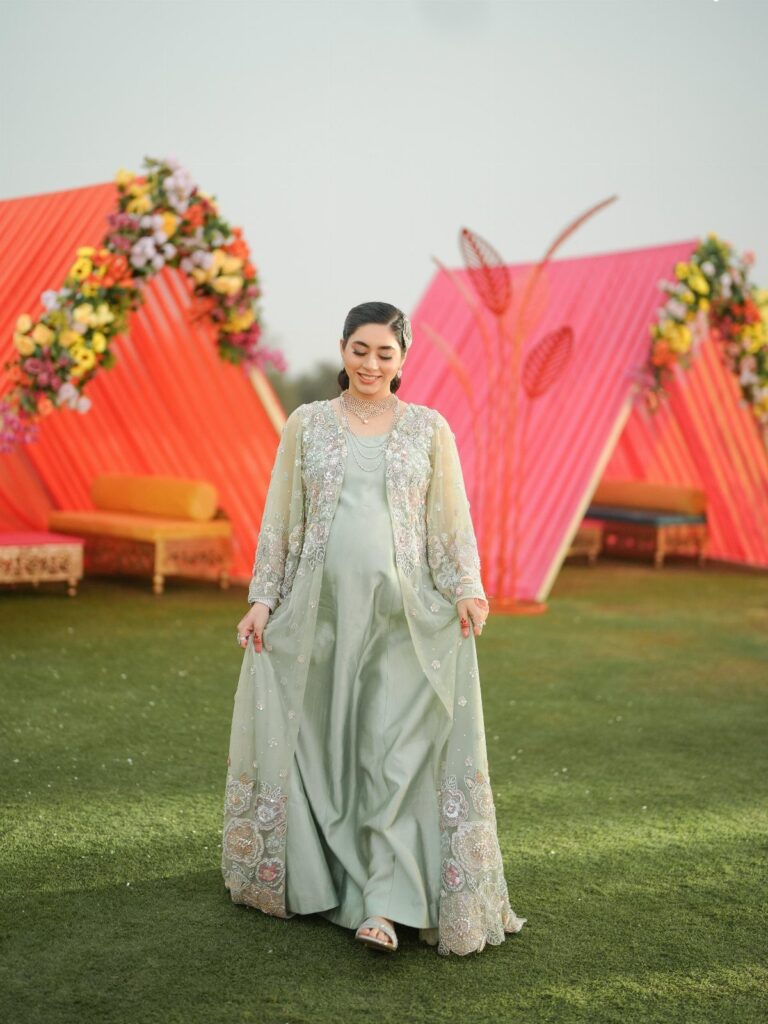Pregnant woman in a light green embroidered gown posing before orange wedding tents.