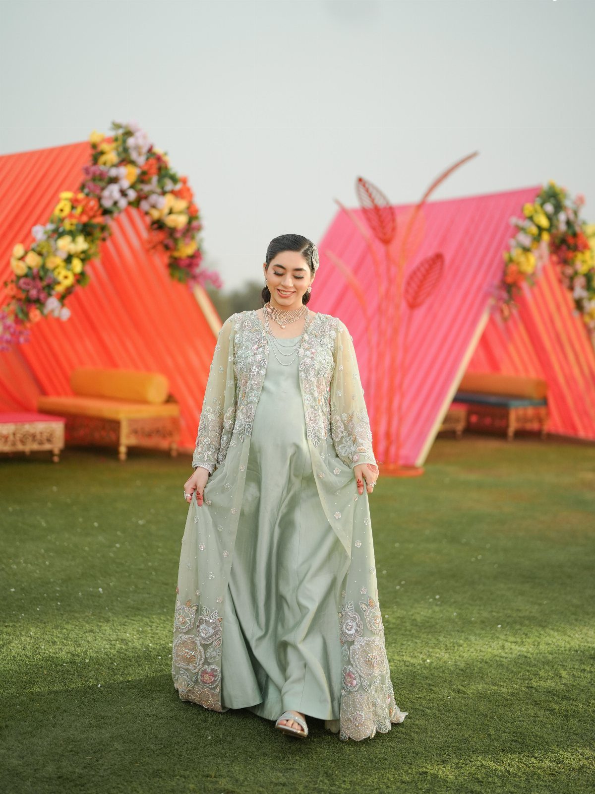 Pregnant woman in a light green embroidered gown posing before orange wedding tents.