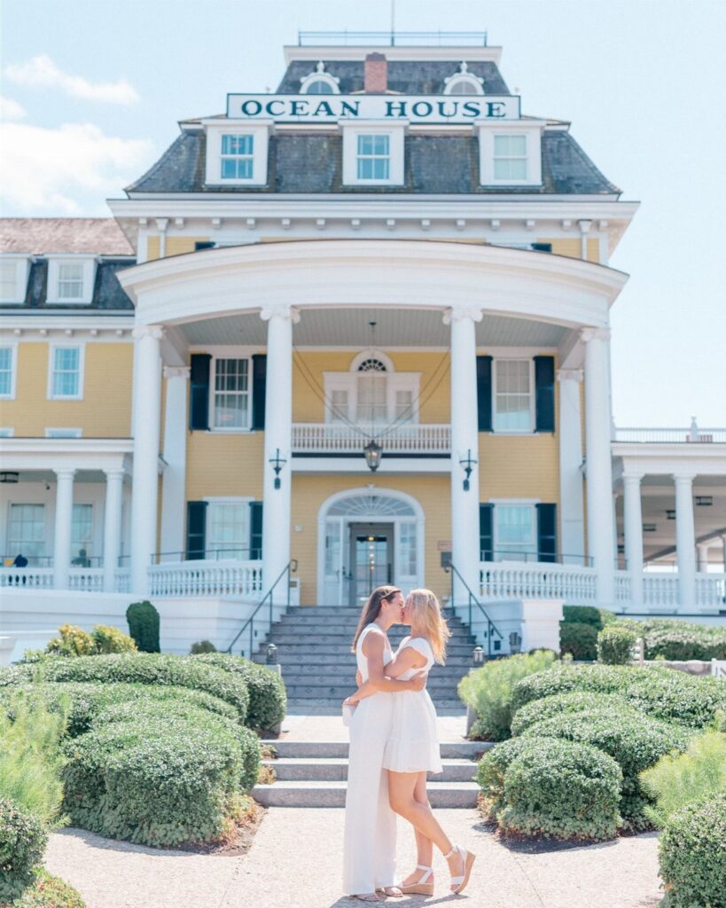 Two women in white dresses kissing before the yellow Ocean House hotel entrance.