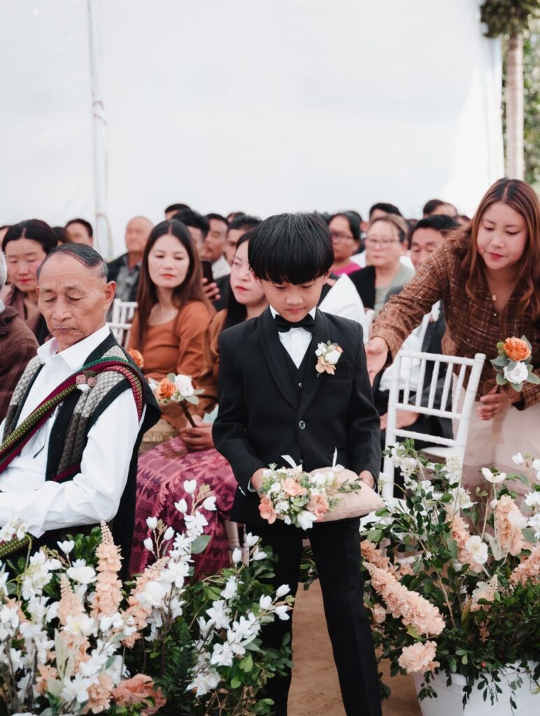 Young boy in a black tuxedo and bow tie holding wedding flowers.