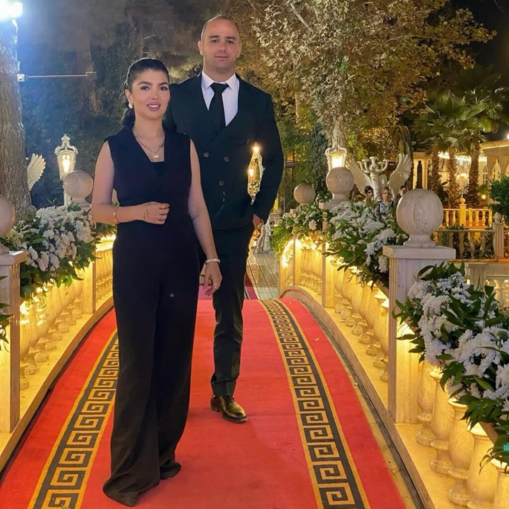 Woman in a black wide-leg jumpsuit and man in a suit on a red carpet.