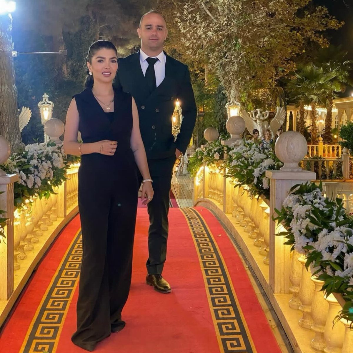 Woman in a black wide-leg jumpsuit and man in a suit on a red carpet.