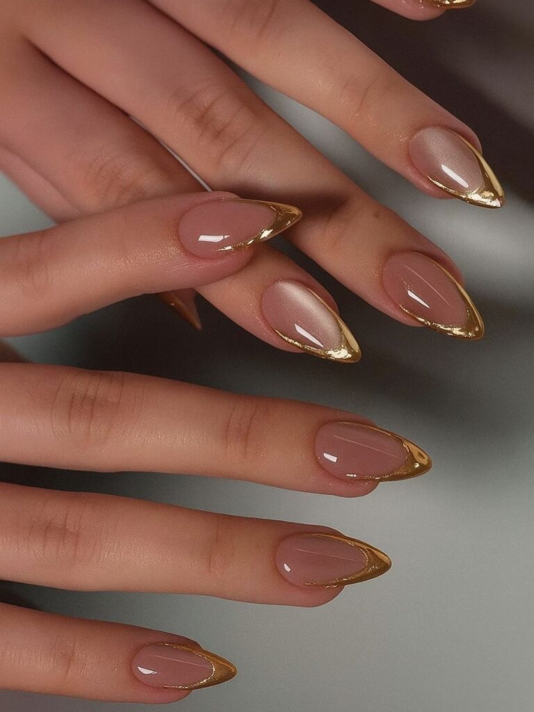 Nude almond-shaped nails with metallic gold French tips on a person's hands.