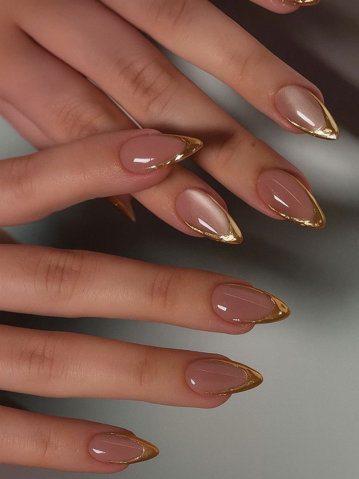 Nude almond-shaped nails with metallic gold French tips on a person's hands.