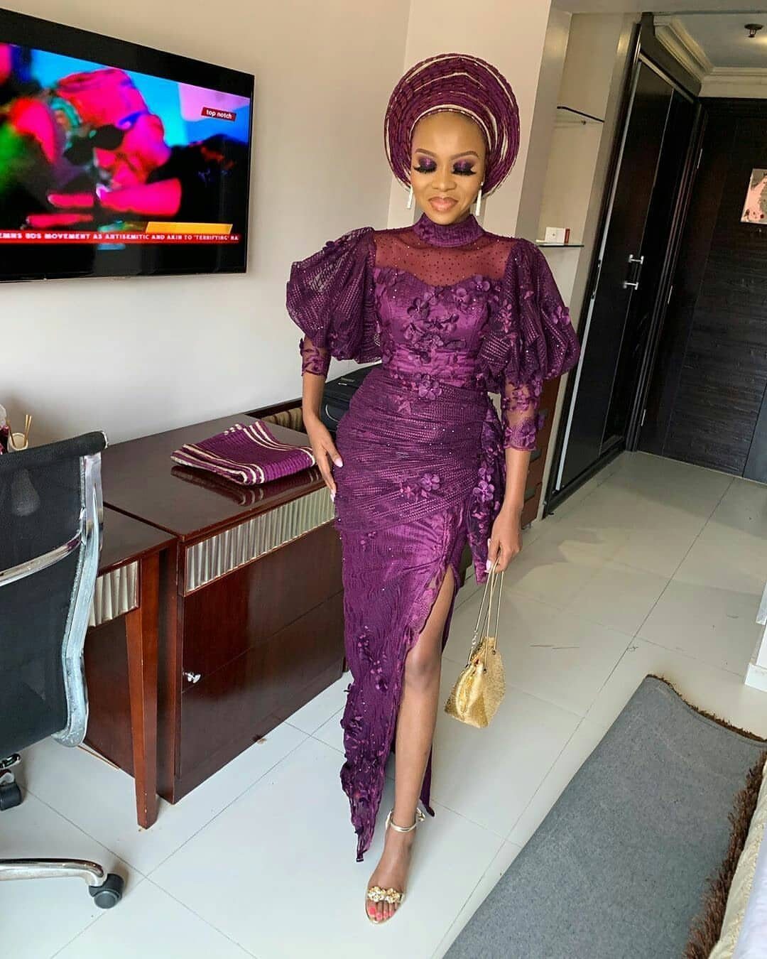 Woman in purple lace gown and matching gele headtie holding a gold clutch.