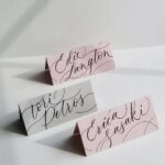Three folded calligraphy place cards in pink and grey on a white background
