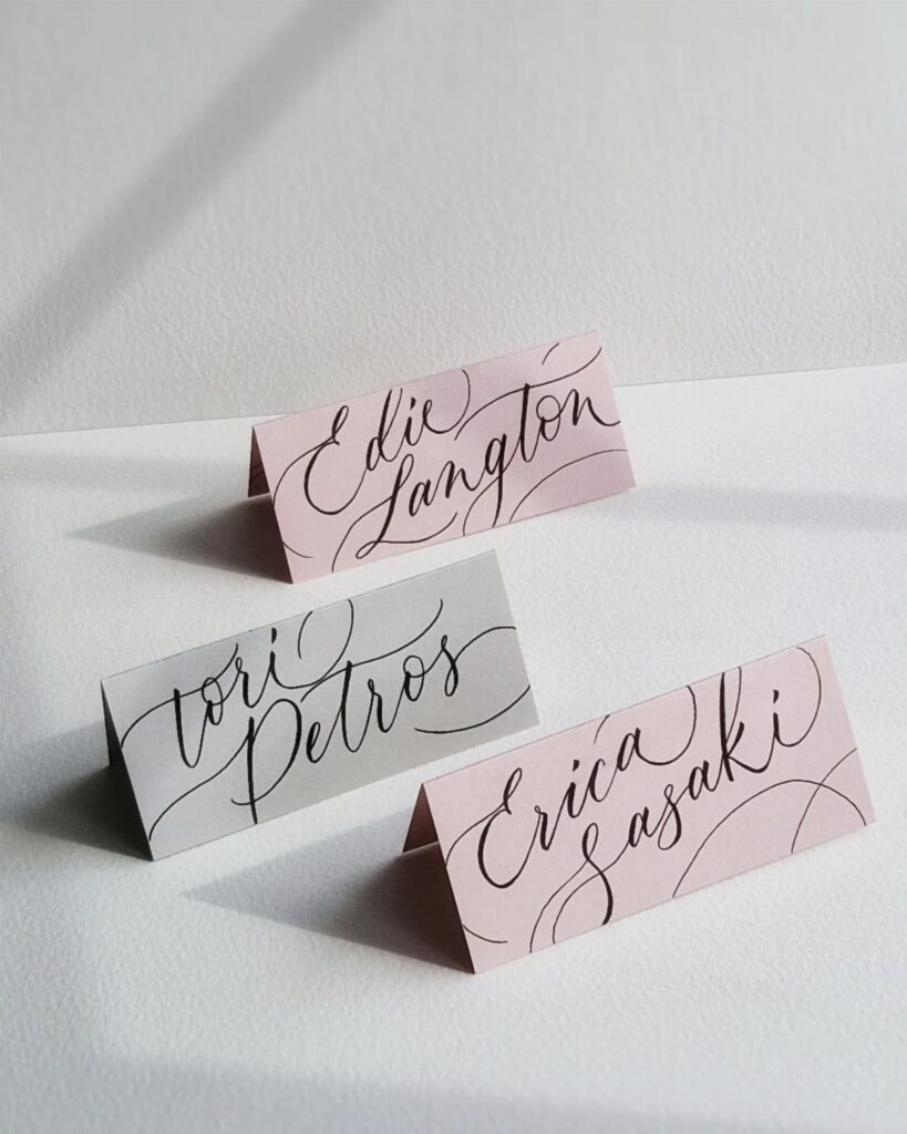 Three folded calligraphy place cards in pink and grey on a white background