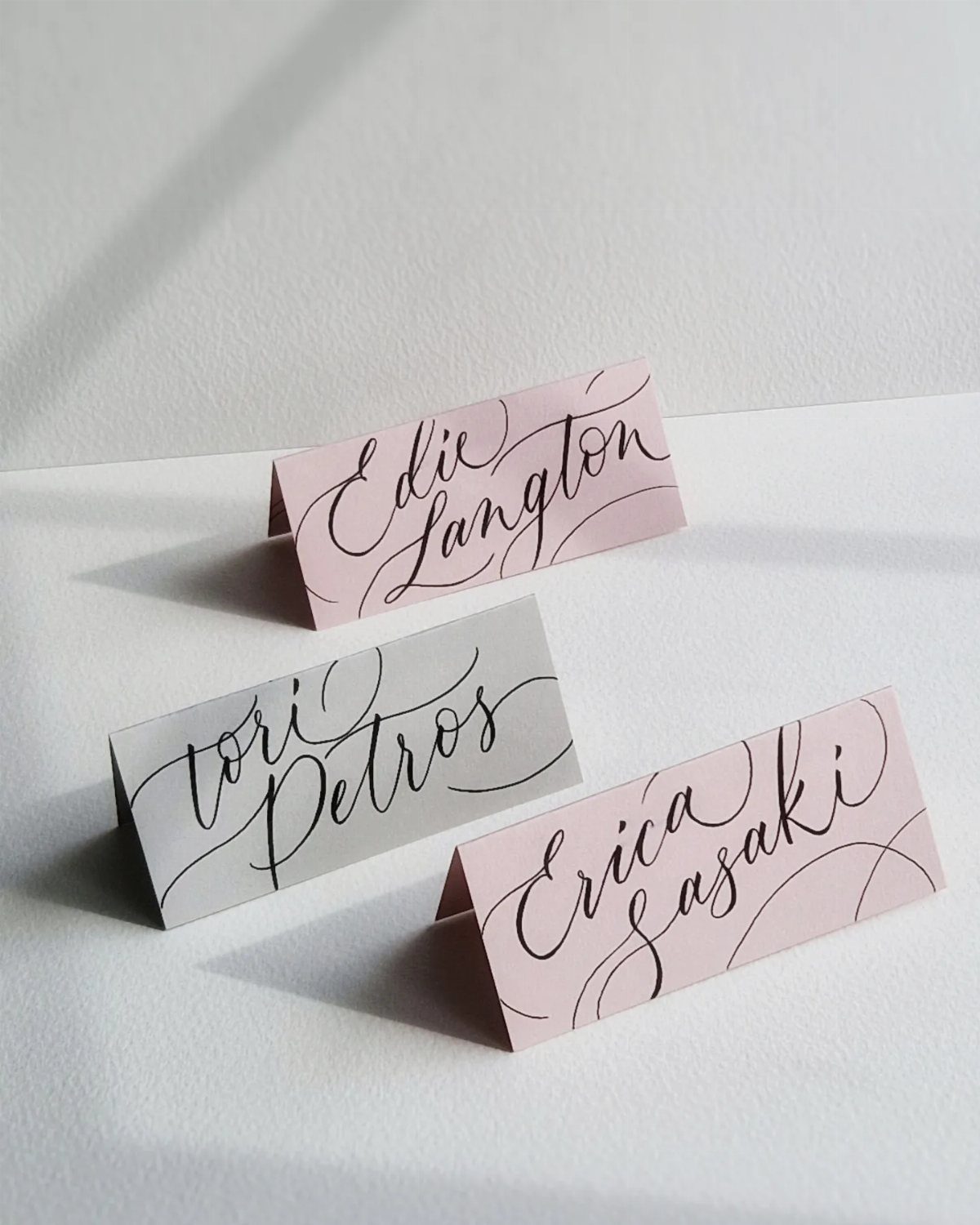 Three folded calligraphy place cards in pink and grey on a white background