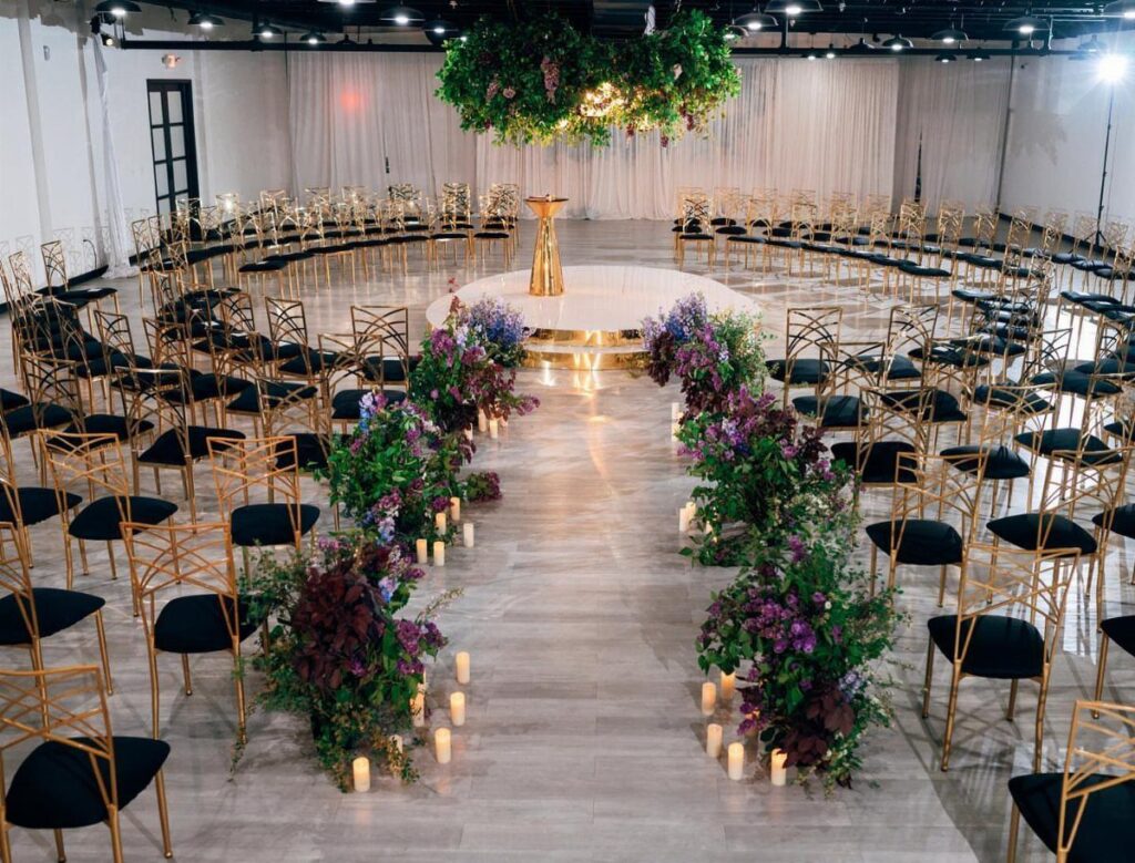 Indoor wedding venue with gold chairs, purple floral aisle arrangements, and hanging greenery.