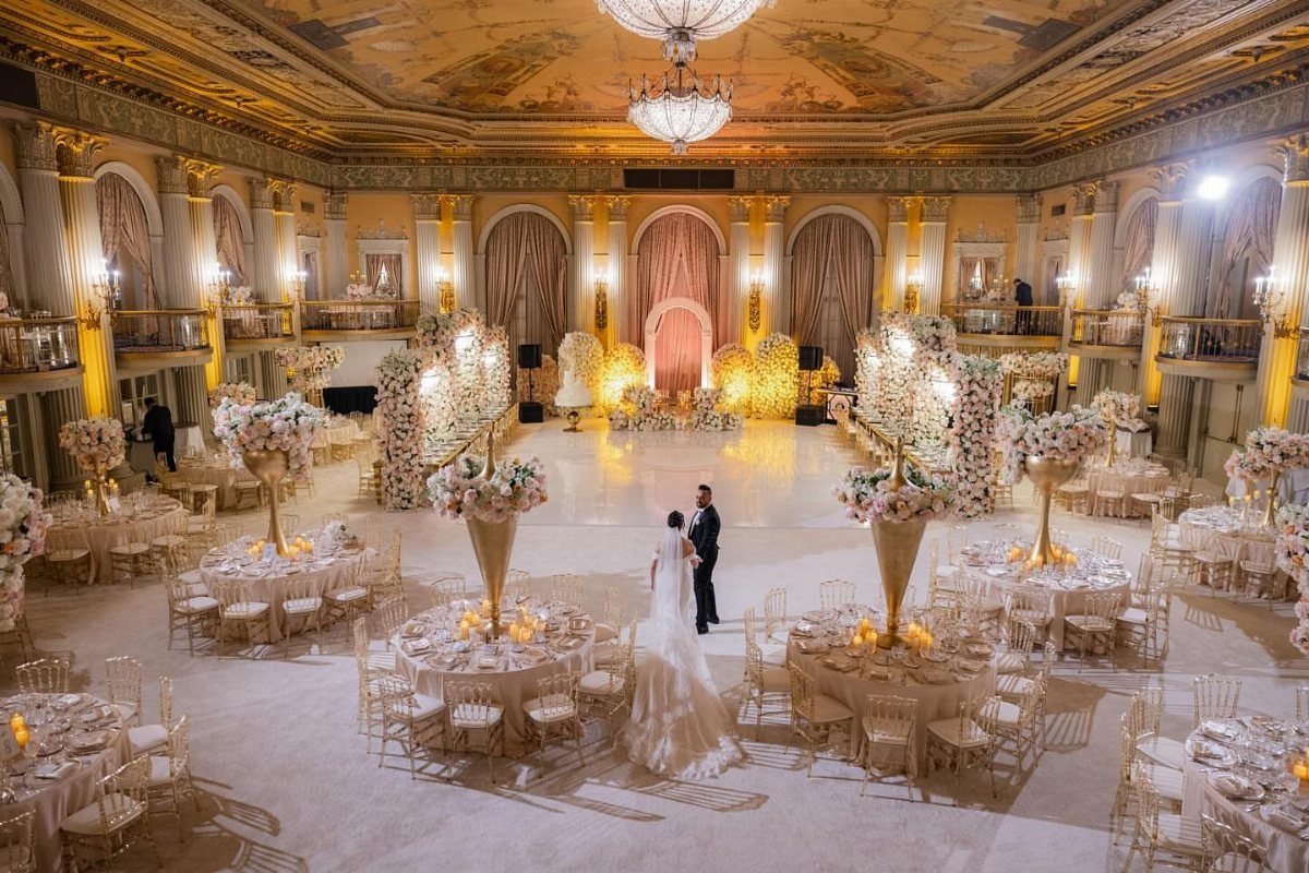Bride and groom in a luxurious gold ballroom with floral centerpieces and chandeliers.