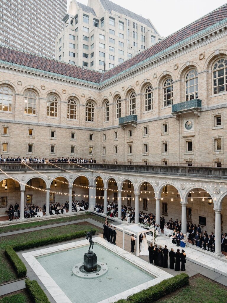 Wedding ceremony in a stone courtyard with arched walkways and a central fountain statue.