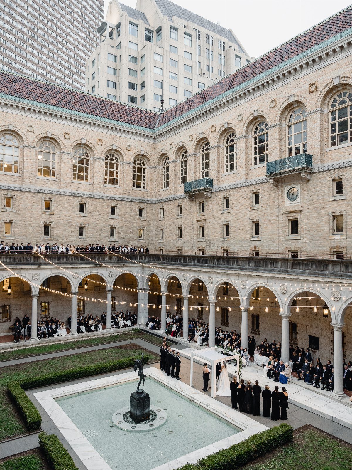 Wedding ceremony in a stone courtyard with arched walkways and a central fountain statue.