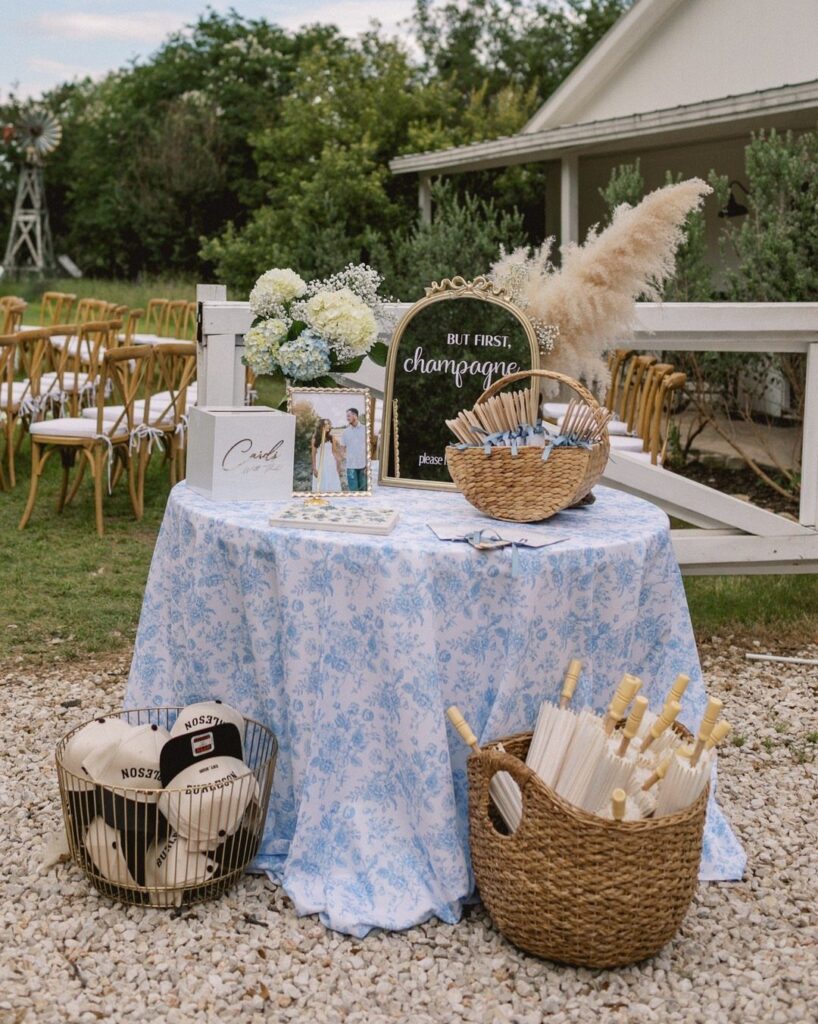 Wedding welcome table with blue floral cloth, champagne mirror, and baskets of drinks.